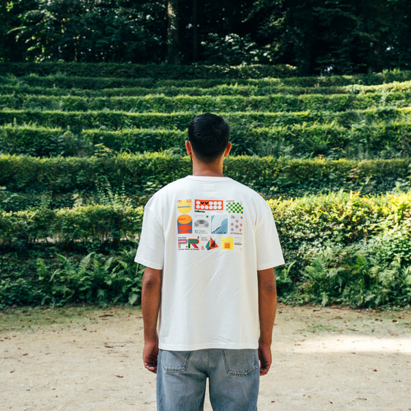 Person wearing a white t-shirt with graphics standing in front of a neatly trimmed hedge maze.